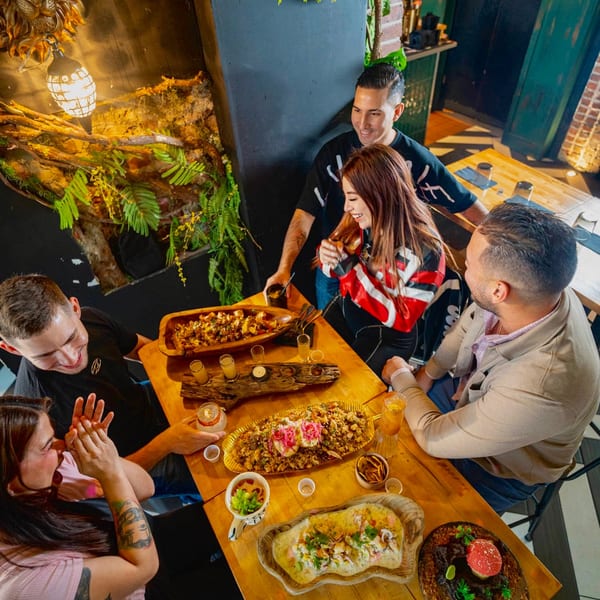 Four friends enjoying a meal with multiple dishes and drinks on a wooden table in a restaurant.