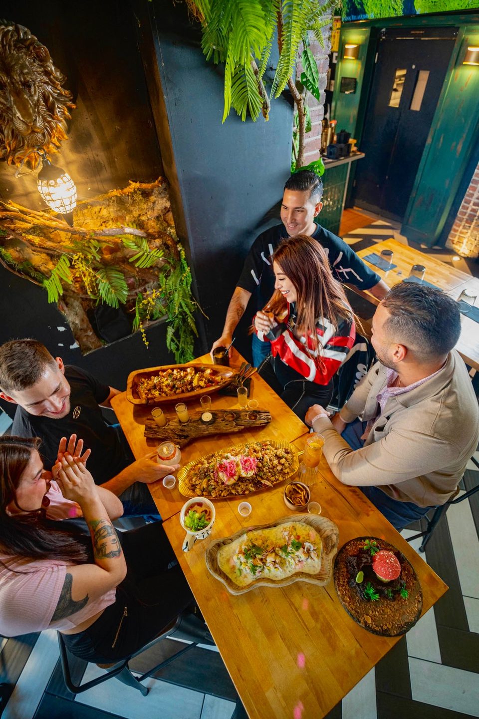 Four friends enjoying a meal with multiple dishes and drinks on a wooden table in a restaurant.