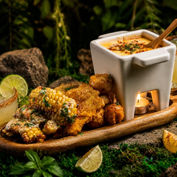 Wooden platter of grilled corn and golden fried bites beside a warm cheese dip, two chili-rimmed lime cocktails, and garnishes.