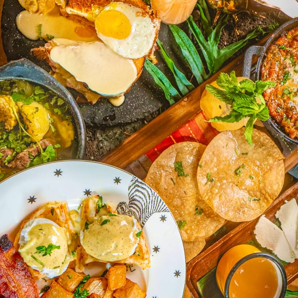 Overhead spread of Latin-inspired dishes, including eggs Benedict, steak, plantains, tortillas, roasted potatoes, sauces, and citrus drinks on a wooden table.