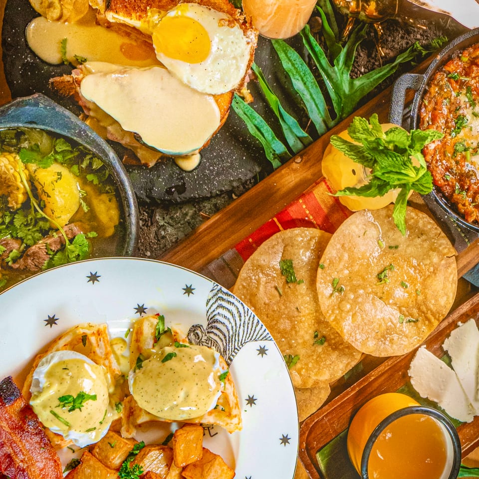 Overhead spread of Latin-inspired dishes, including eggs Benedict, steak, plantains, tortillas, roasted potatoes, sauces, and citrus drinks on a wooden table.