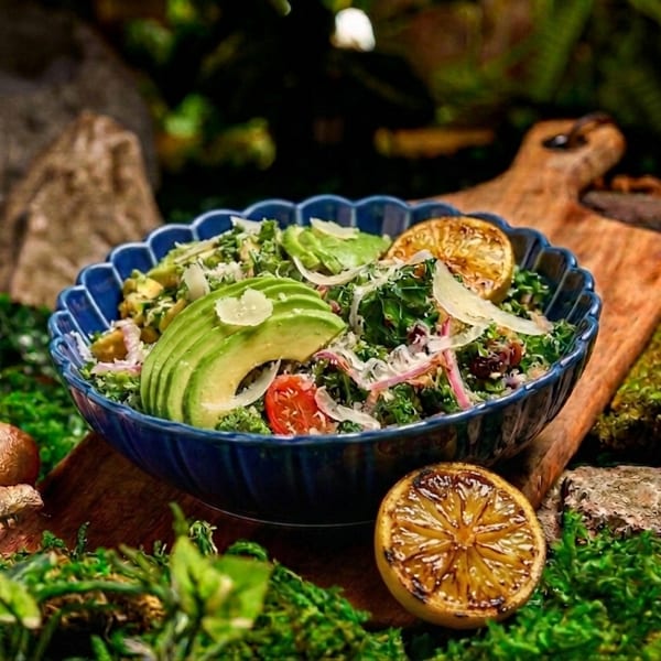 Colorful salad with avocado, greens, tomatoes, onions, and citrus sits in a blue bowl on a rustic wooden board surrounded by moss.